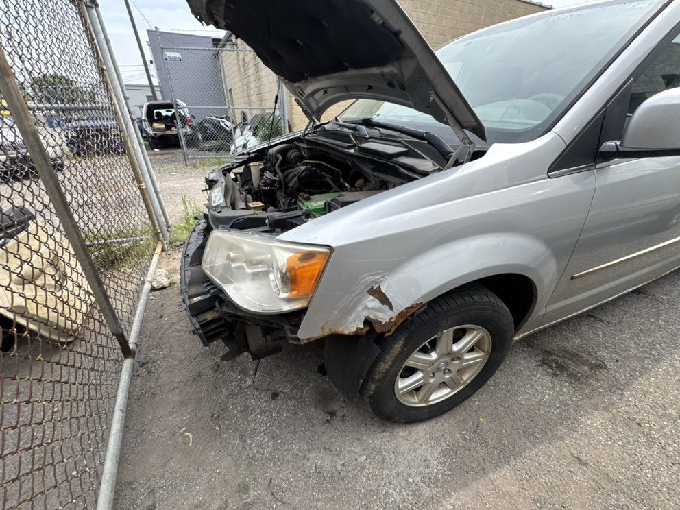 Silver minivan with front-end damage, hood open, parked next to a chain-link fence.