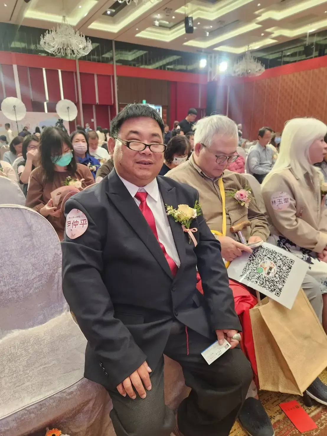 活動現場,一位身穿黑色西裝、繫著紅色領帶的男士面帶微笑。他坐在室內,背景是觀眾。