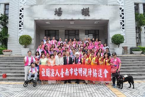 一群人在建築物外合影,手舉橫幅。許多人穿著粉紅色和黃色服裝。現場有一隻狗和一輛輪椅。