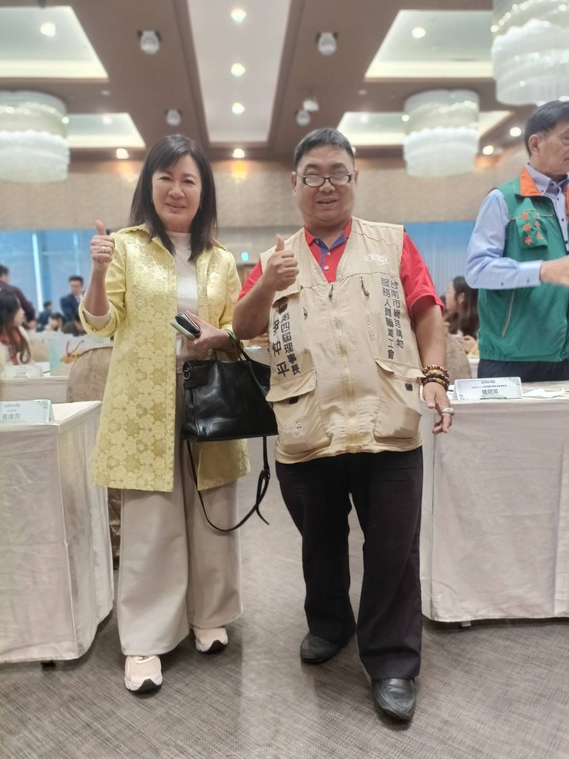 兩個人豎起大拇指。一位身穿黃色外套和褲子的女士,一位身穿背心的男士,在一場會議上。