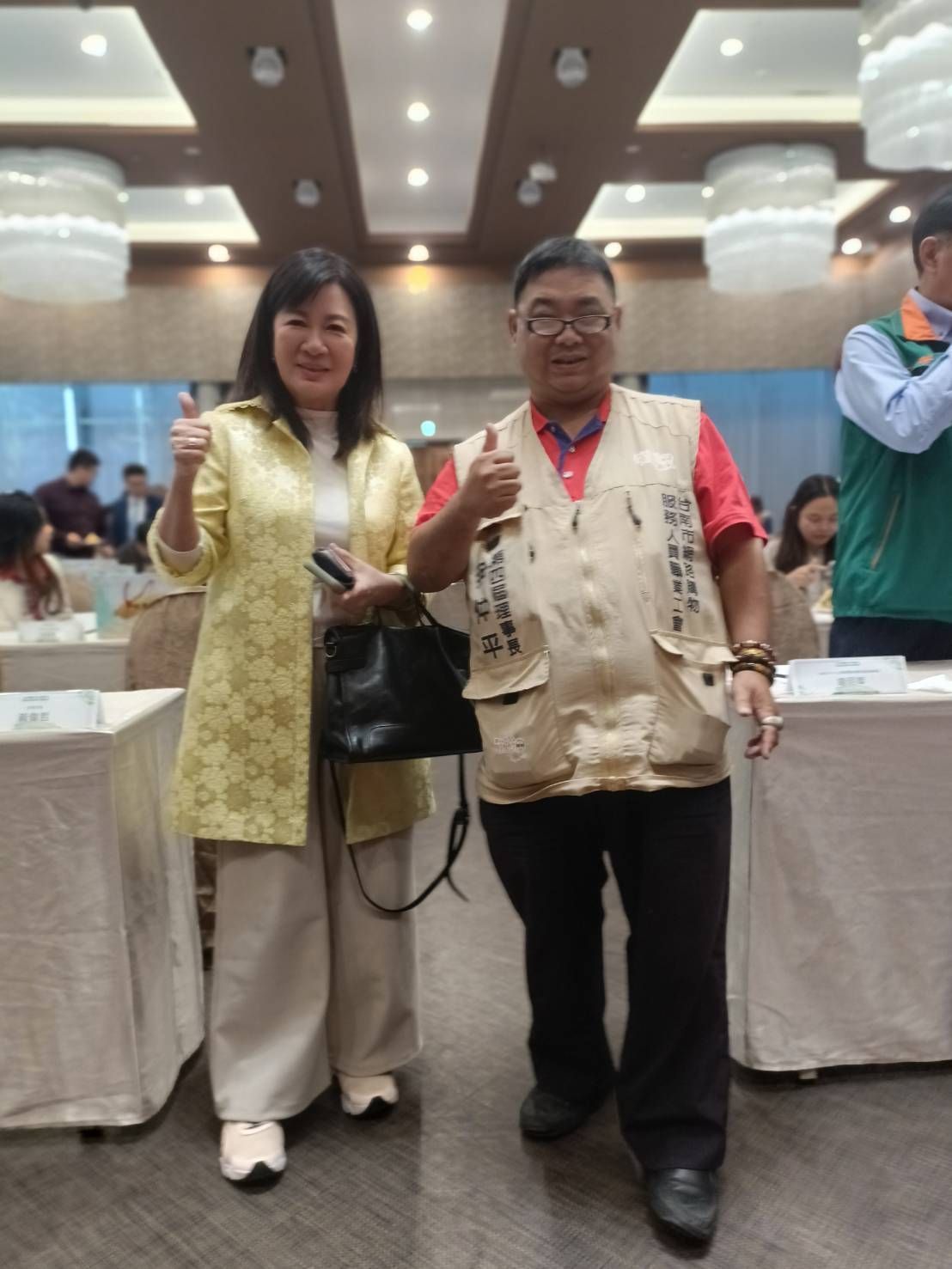 兩個人豎起大拇指。會議室裡,一位女士身穿黃色夾克,一位男士身穿背心。