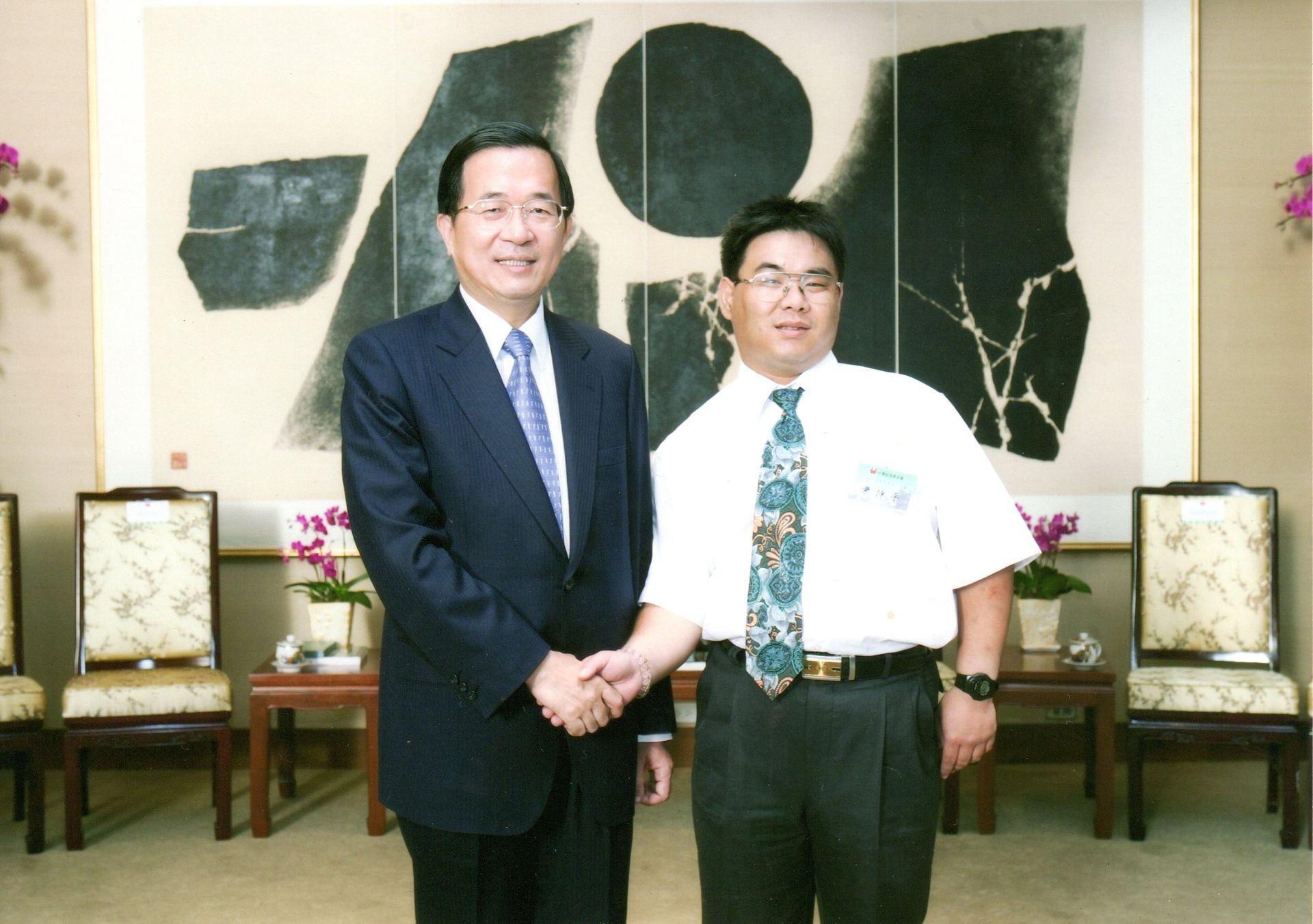 兩位男士在一幅大型抽象畫前握手。一位身著西裝,另一位則穿著休閒襯衫,打著領帶。