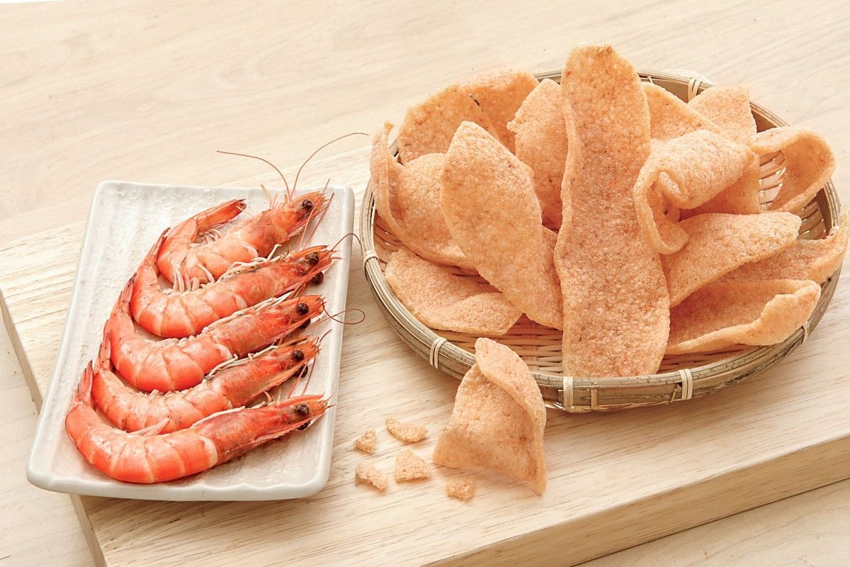 木質表面上，一籃蝦片旁邊放著煮熟的蝦子。