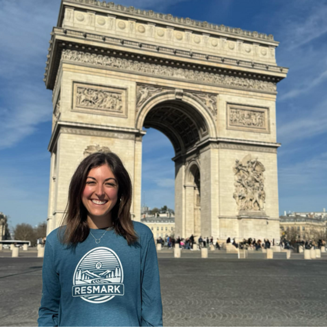 Woman with brown hair smiles, wearing gray and orange shirt in front of a blurred green forest.