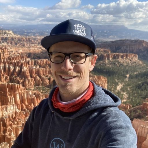 Man in a blue cap and glasses smiles, standing in front of a canyon with red rock formations.