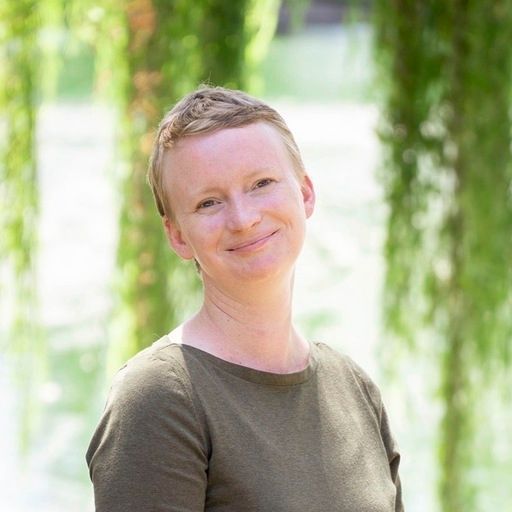 Woman with short blonde hair smiles outdoors; wearing a green shirt, blurred green foliage backdrop.