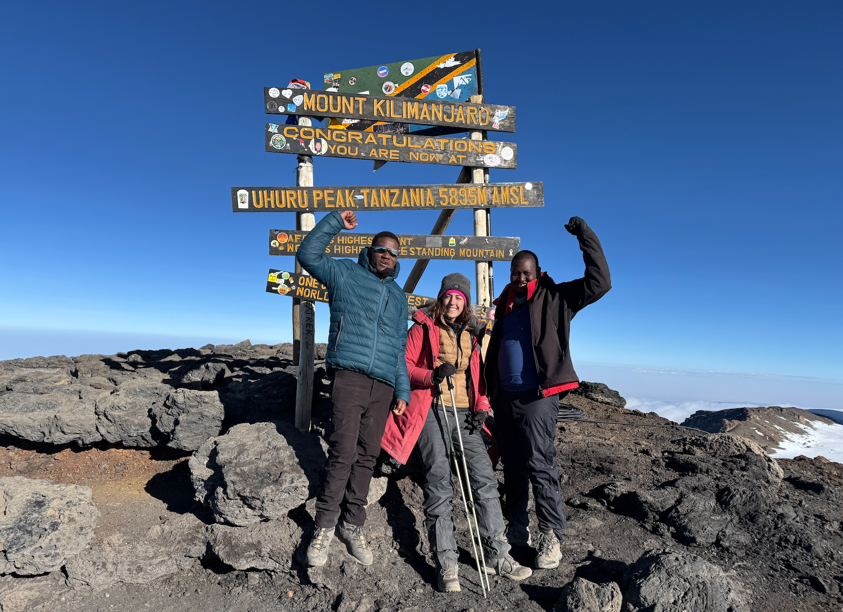 Three people celebrate on Mount Kilimanjaro summit. Sign post indicates peak. Blue sky.