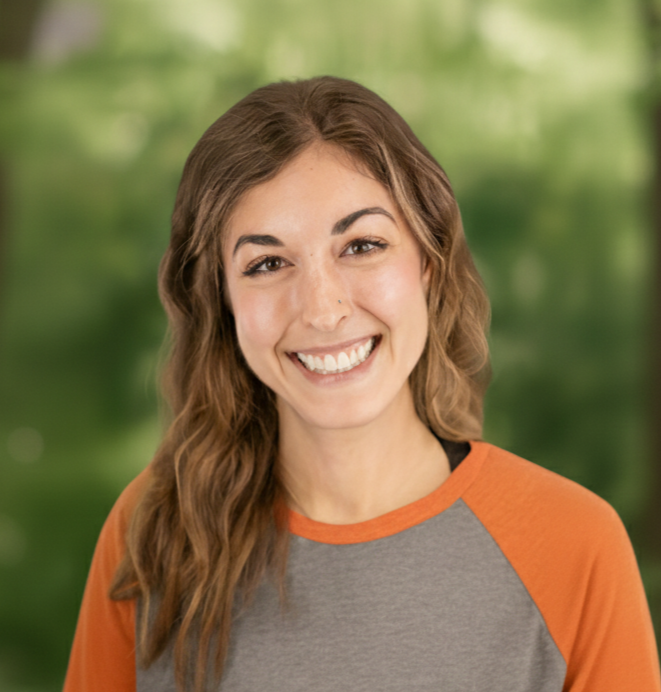 Woman with brown hair smiles, wearing gray and orange shirt in front of a blurred green forest.