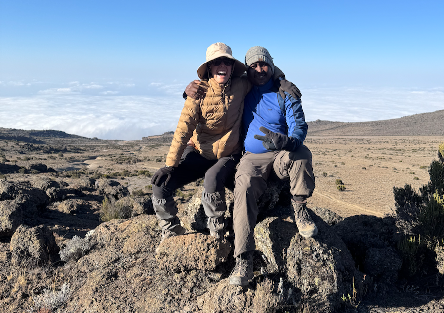 Two people sit on a rocky mountaintop, arms around each other, above a sea of clouds under a blue sky.