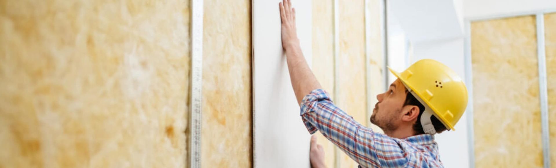 A man wearing a hard hat is measuring a wall with a tape measure.