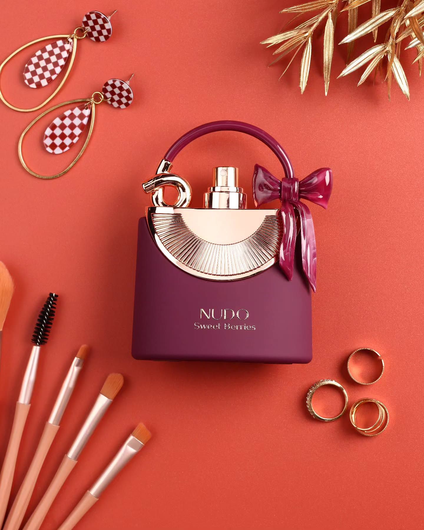 Frasco de perfume morado con lazo y asa, pinceles de maquillaje, pendientes y anillos dorados sobre fondo naranja.