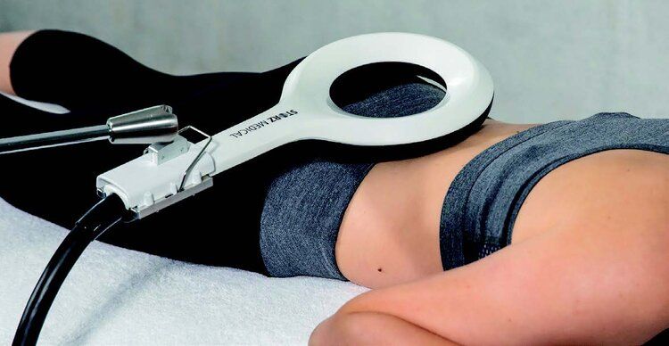 A person lying face down receiving magnetic stimulation on their lower back from a circular device.