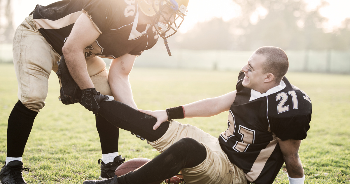 Picture of injured player Football player on the ground, holding his leg, being assisted by a teammate. Both wear black and gold uniforms on a grassy field.