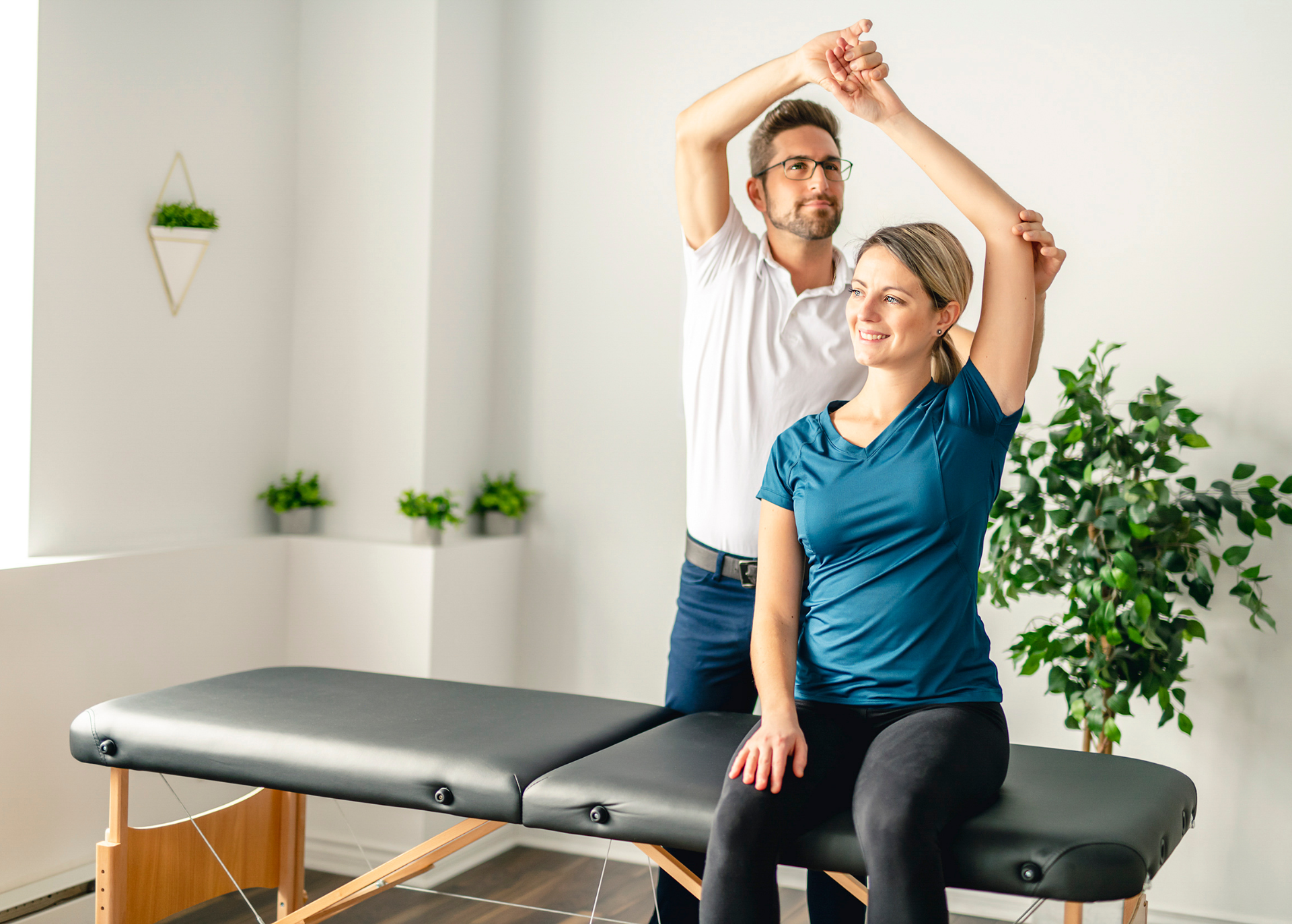 Shoulder testing by physiotherapist Physical therapist assisting a woman with arm movement in a well-lit room. The woman smiles while seated on a massage table.