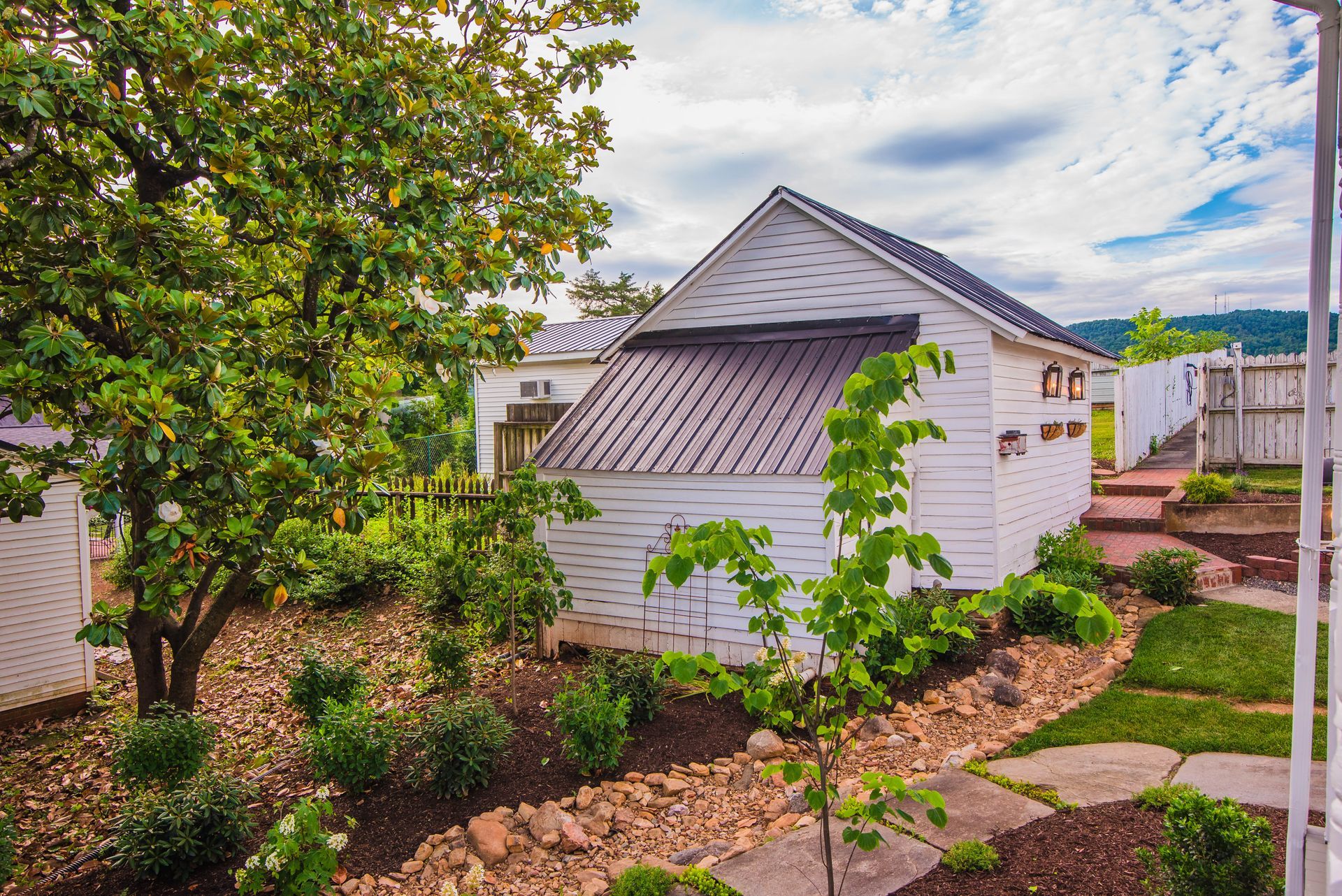 A white shed with a brown roof is surrounded by trees and bushes in a backyard.
