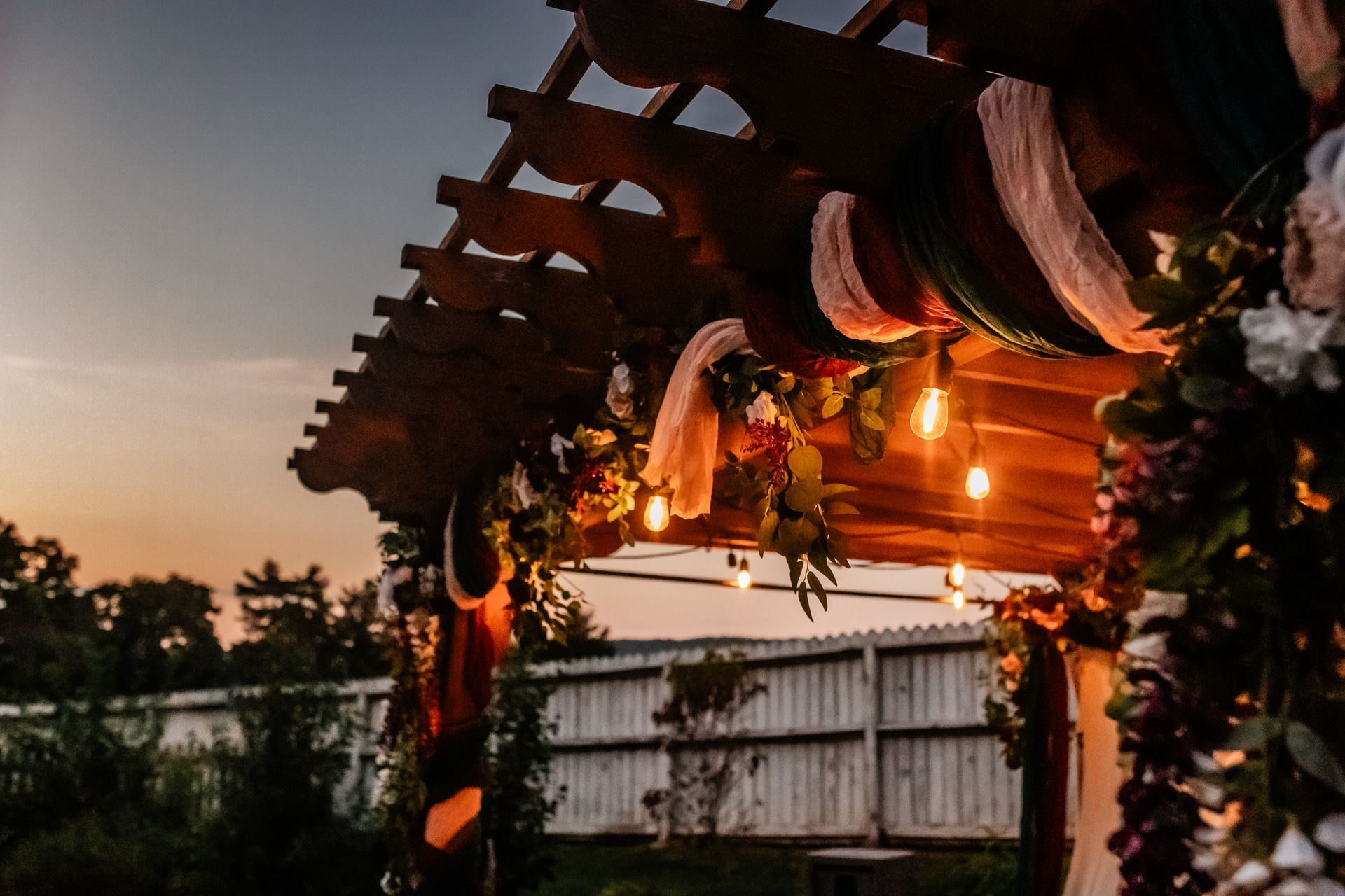 A wooden pergola decorated with flowers and lights at sunset.