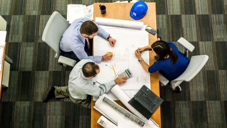 Three professionals sit at a desk reviewing architectural blueprints in an office with gray carpet.