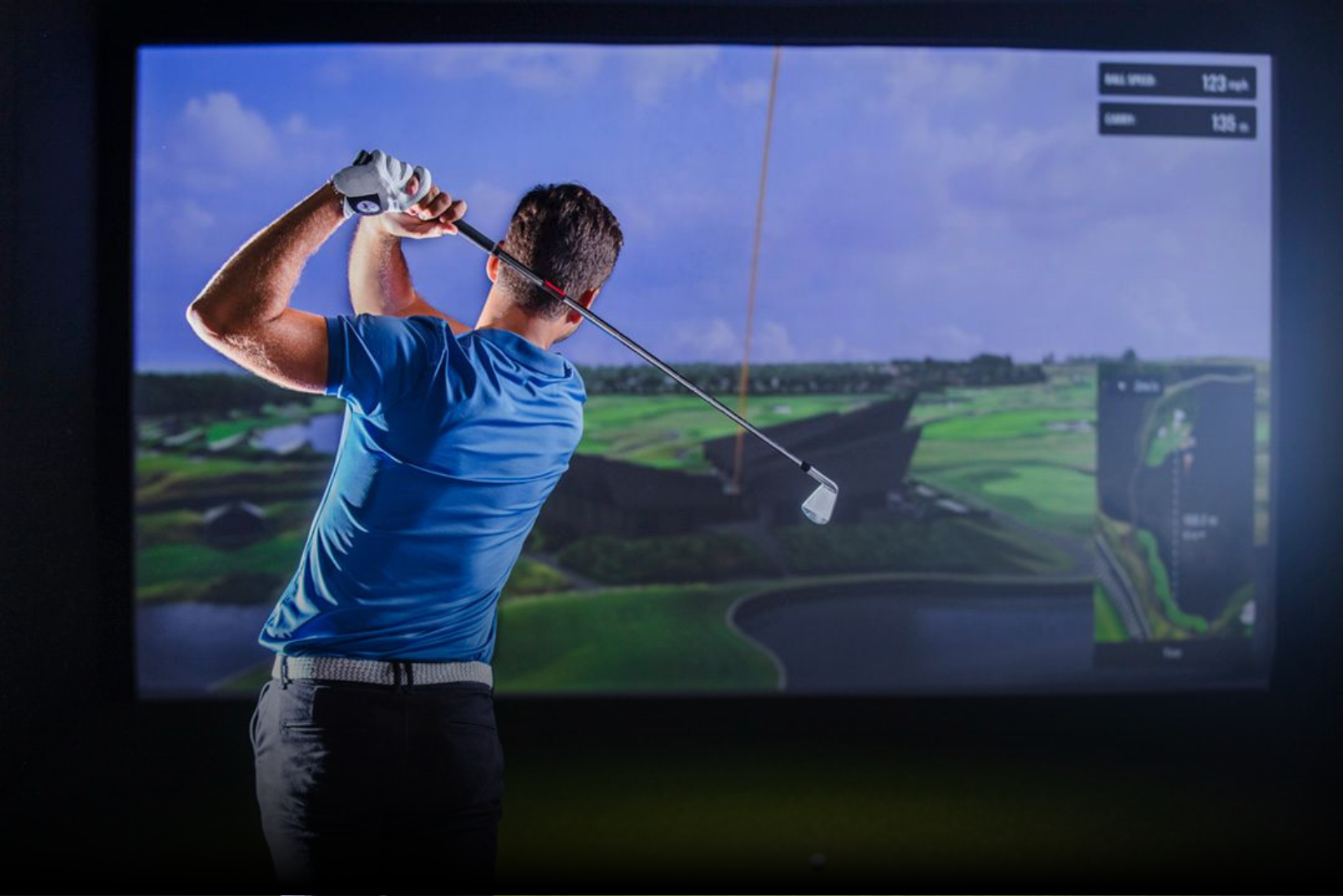 A man is swinging a golf club in front of a large screen.