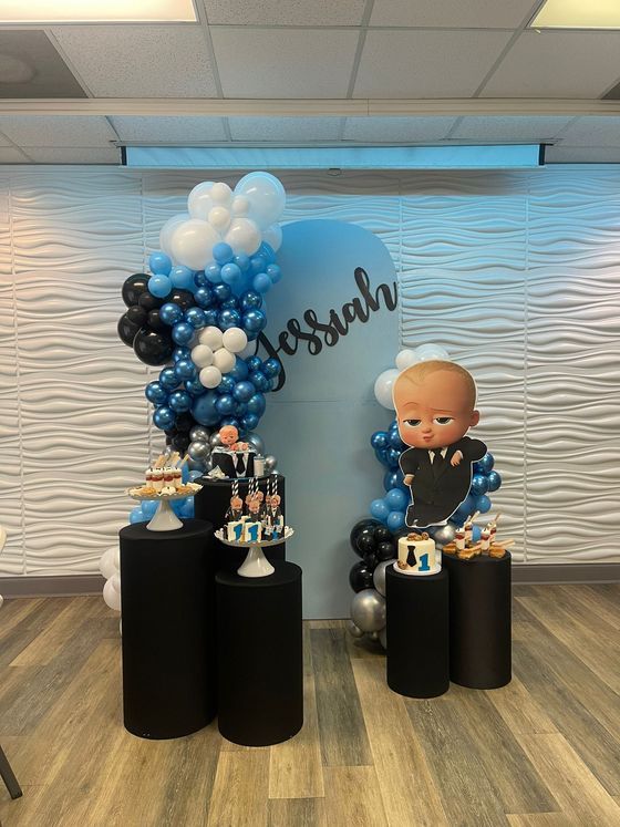 Boss Baby-themed birthday party: balloon arch, cakes, black pedestals, Boss Baby figure. Light blue background.