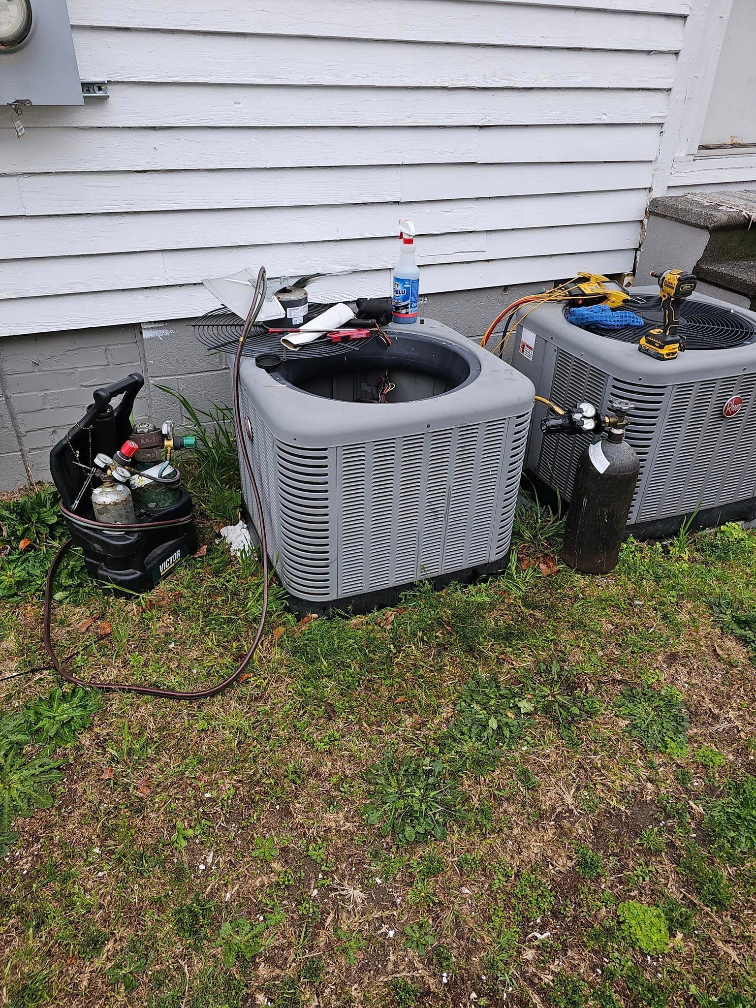 Two air conditioners are sitting in the grass in front of a house.