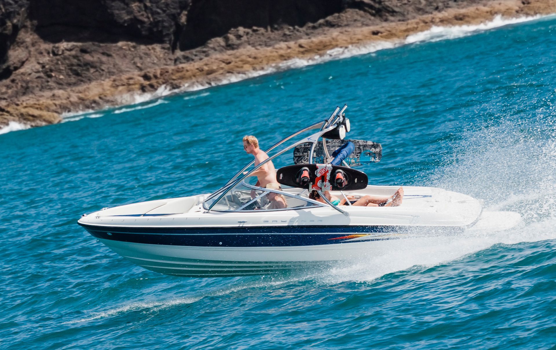 Boat speeding on ocean with people, tow rope, and wakeboard near rocky shore.