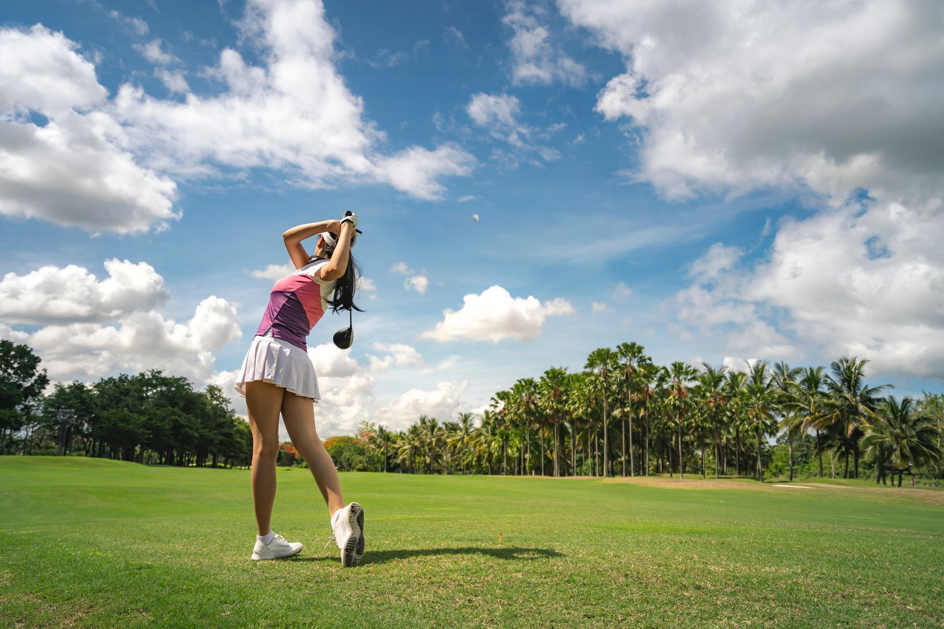 Woman in golf attire swinging a club on a green golf course under a blue sky with fluffy clouds.