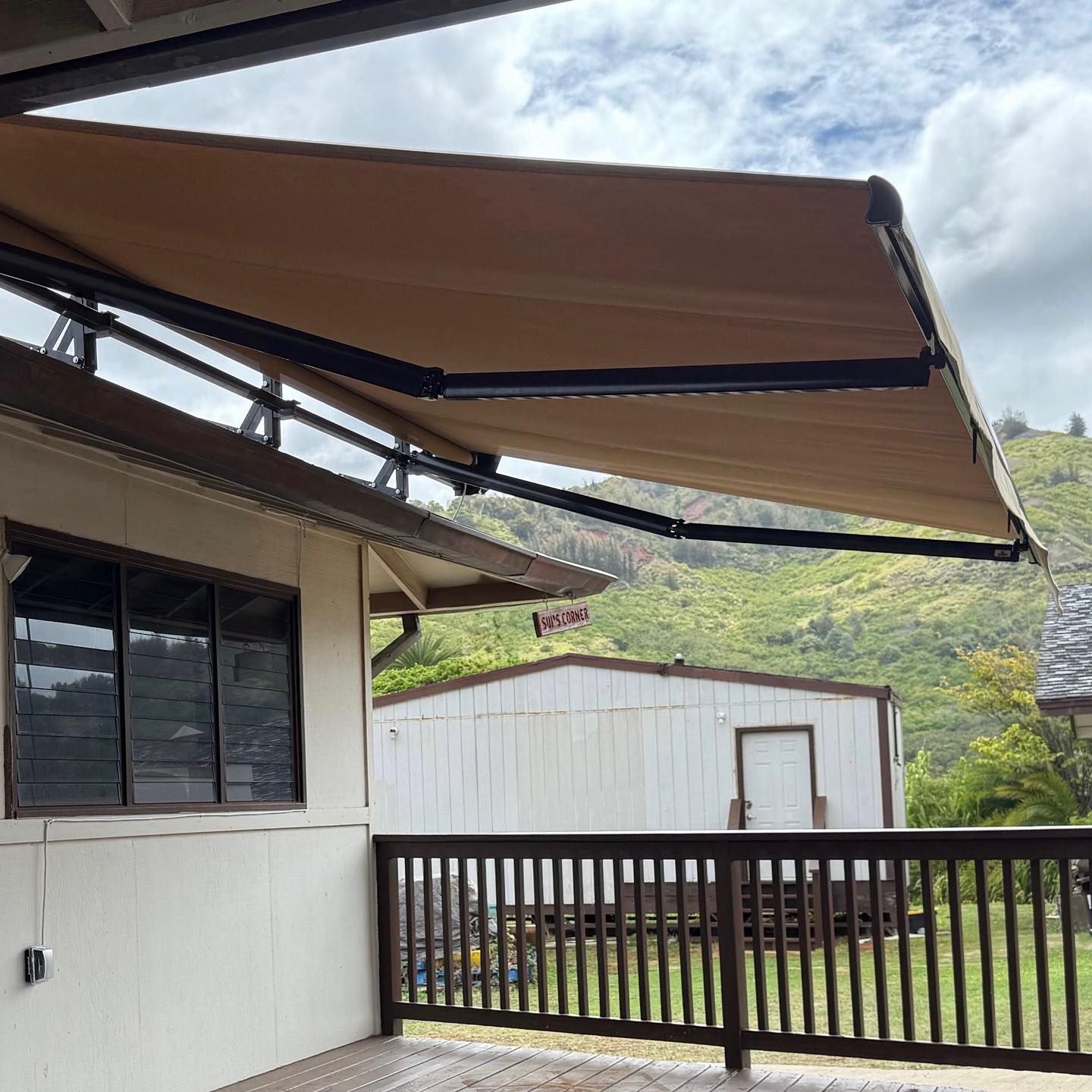 Beige awning over a patio beside a house, with a white shed, black railing, and green hills under cloudy sky.