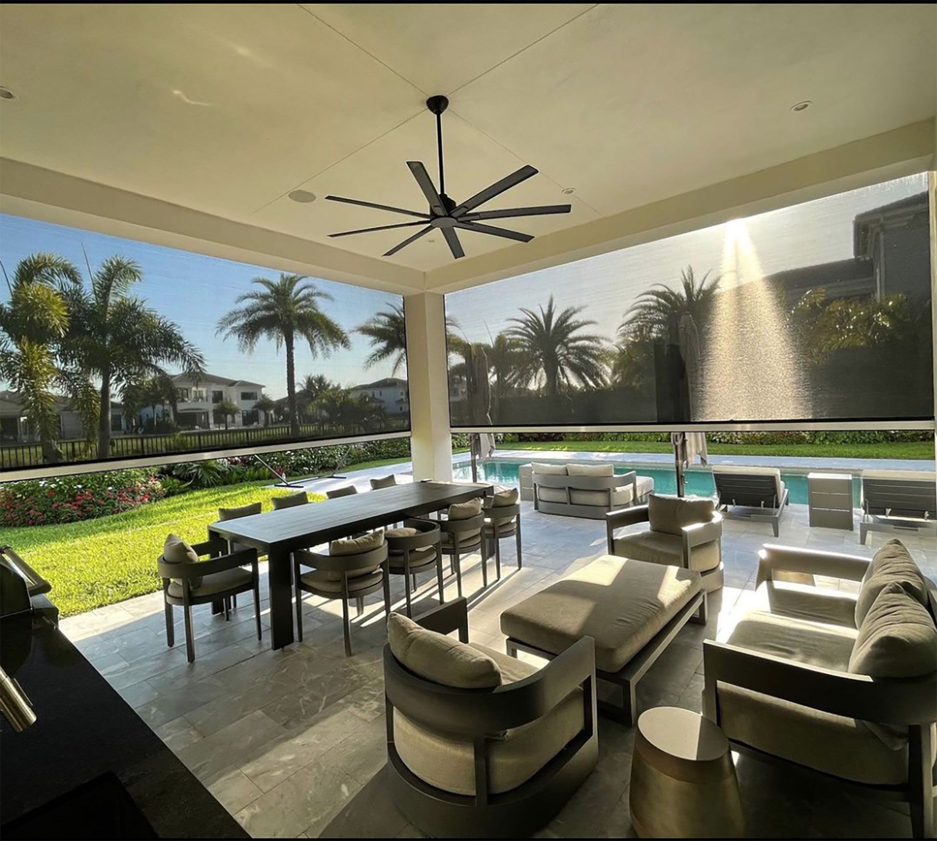Covered patio lounge with seating, dining table, pool, palm trees, and sunset light in a tropical backyard