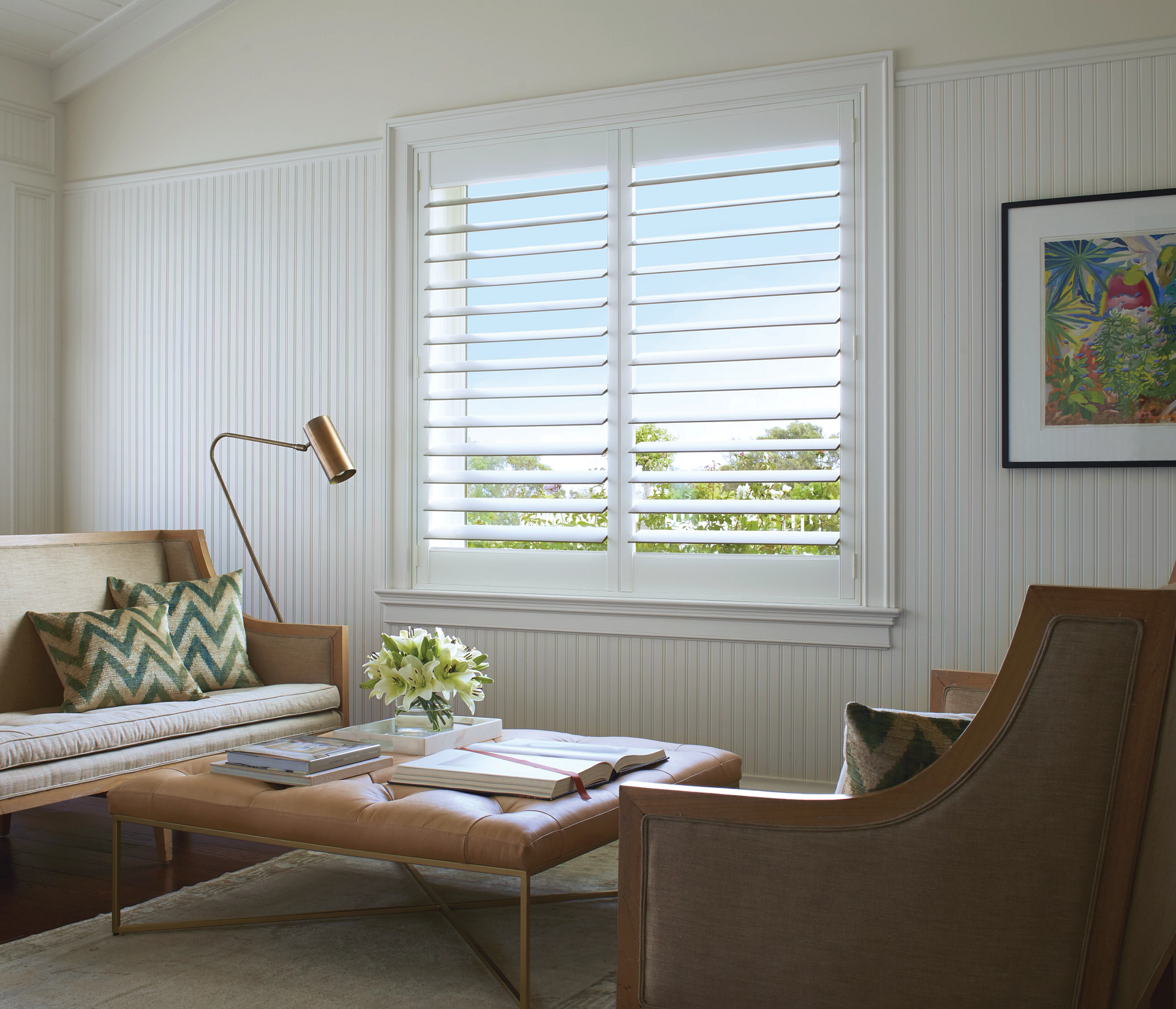 Bright living room with white shutters, beige sofa, wood coffee table, and wicker chair by a window.