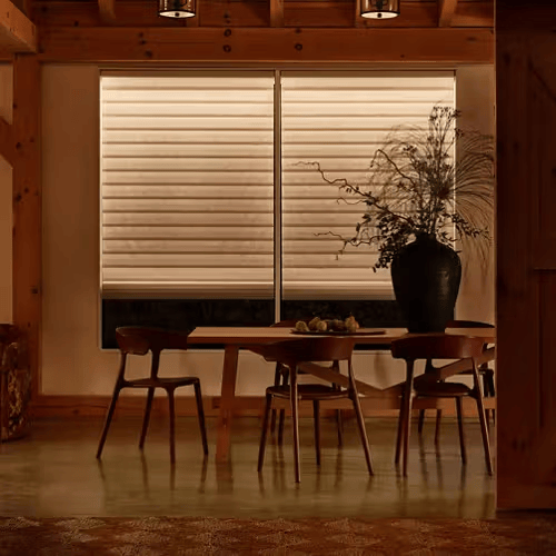 Warm dining room with a wooden table, chairs, and a large vase beside closed blinds.