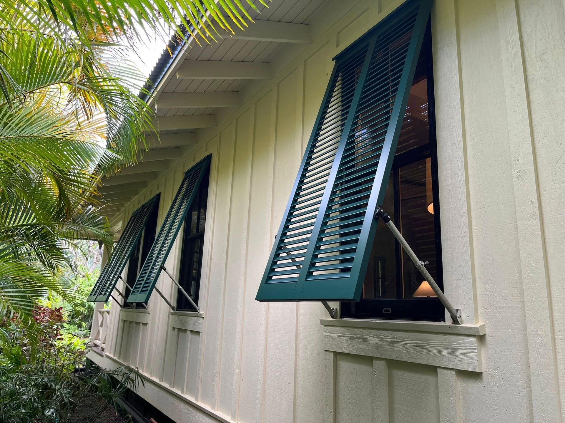 Open green shutters on a white house exterior with tropical plants nearby