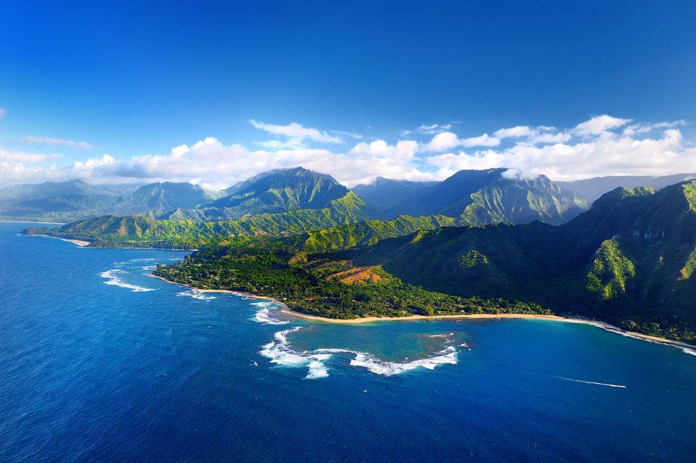 Aerial view of a lush tropical coastline with blue ocean, green cliffs, and a sandy beach under a clear sky