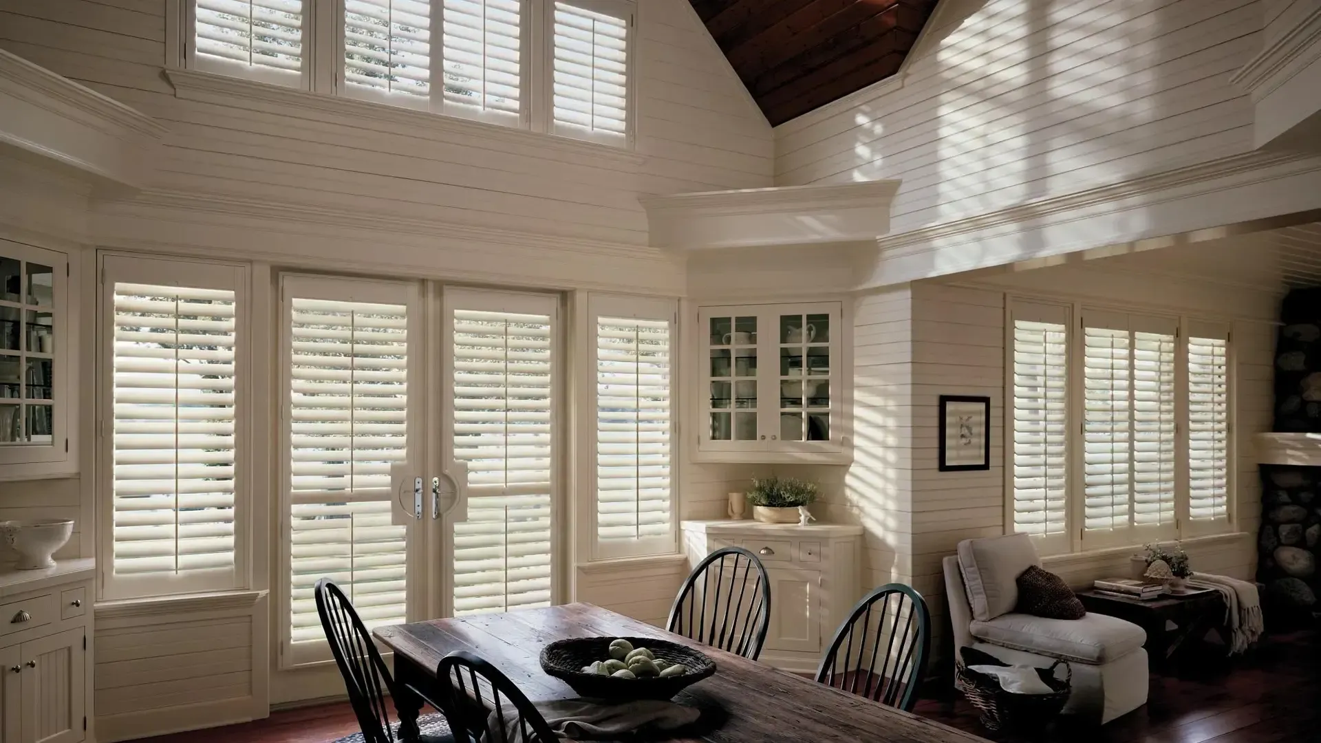 Sunlit living room with white shutters, dining table, and beige sofa beneath a vaulted wood ceiling