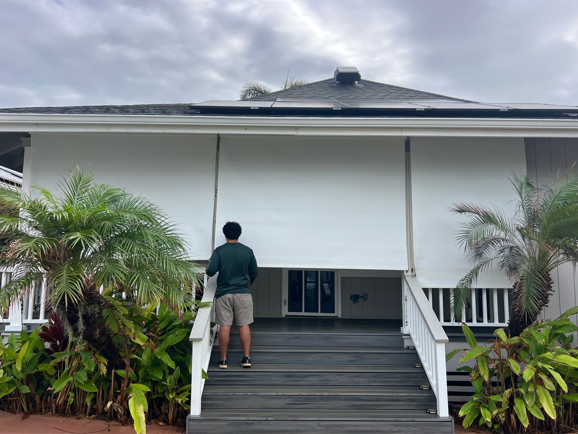 Person standing on steps of a white house with a dark roof and tropical plants on both sides