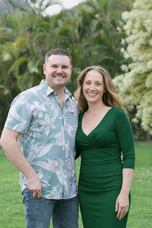 Smiling couple standing outdoors in front of lush greenery, wearing green and blue floral outfits