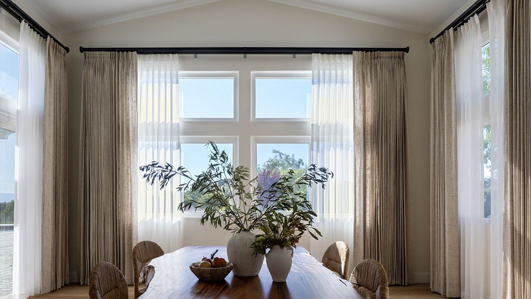 Bright dining room with a wooden table, potted plants, and large windows with sheer curtains