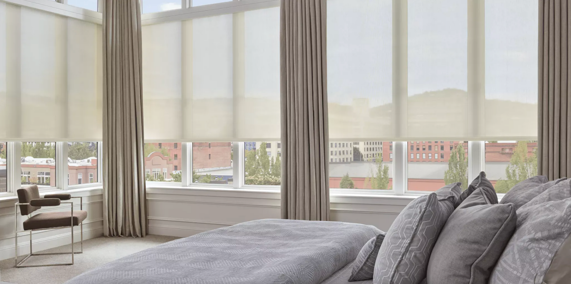 Bright bedroom with large windows, beige curtains, and a bed with gray pillows in the foreground