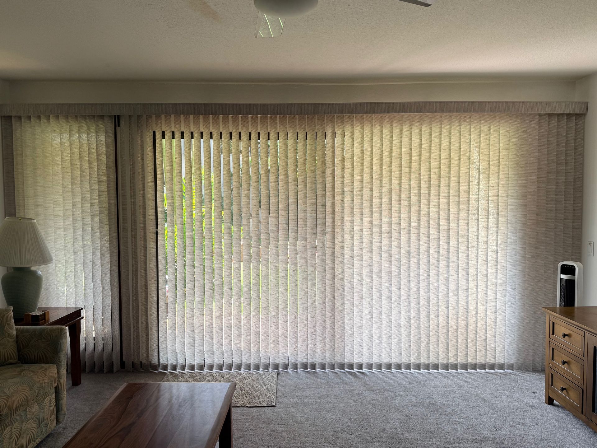 Living room with vertical blinds covering a bright sliding glass door, flanked by a sofa and wooden cabinet.