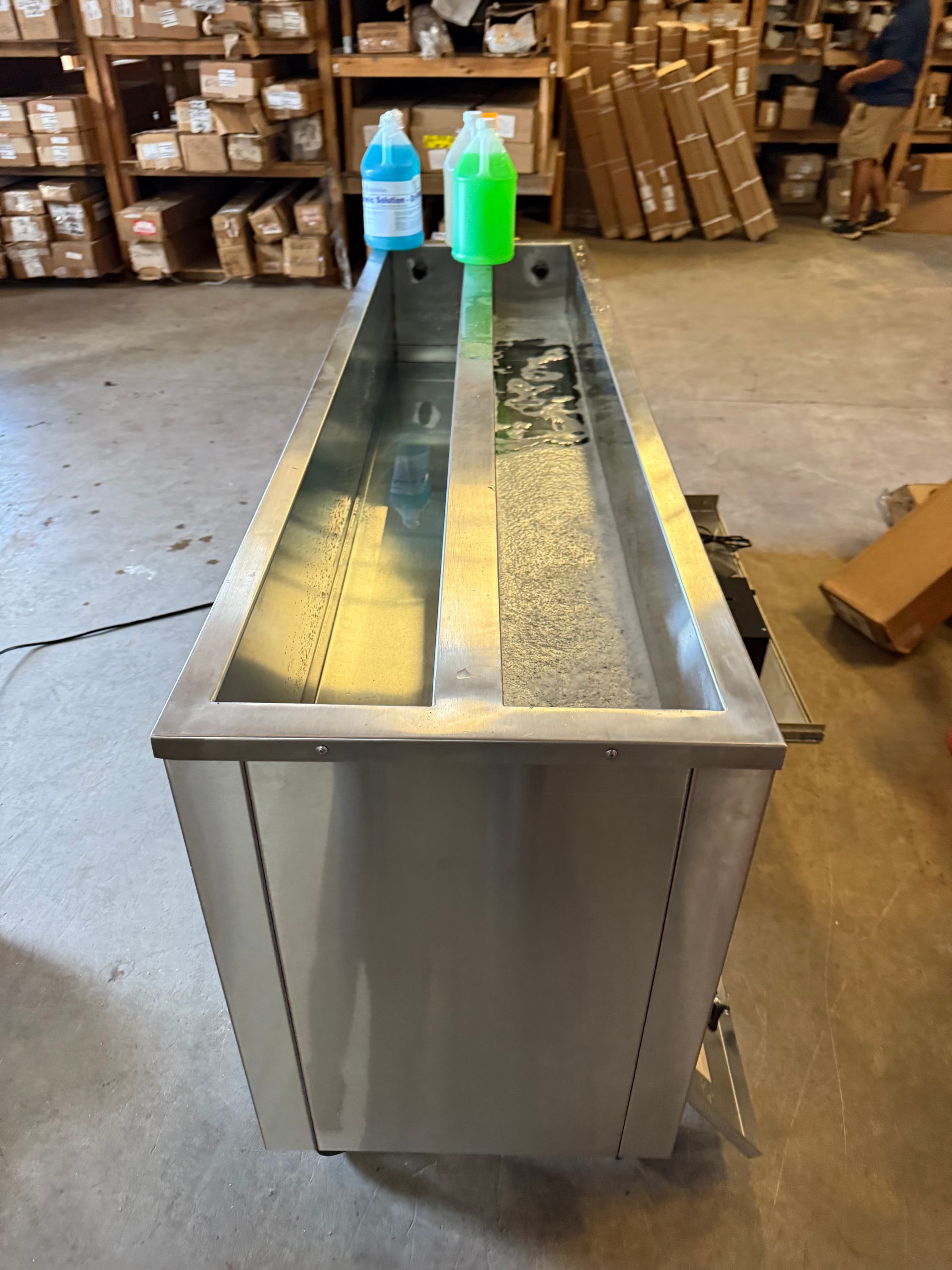 Stainless steel trough with two bottles on top in a warehouse aisle, with stacked boxes in the background