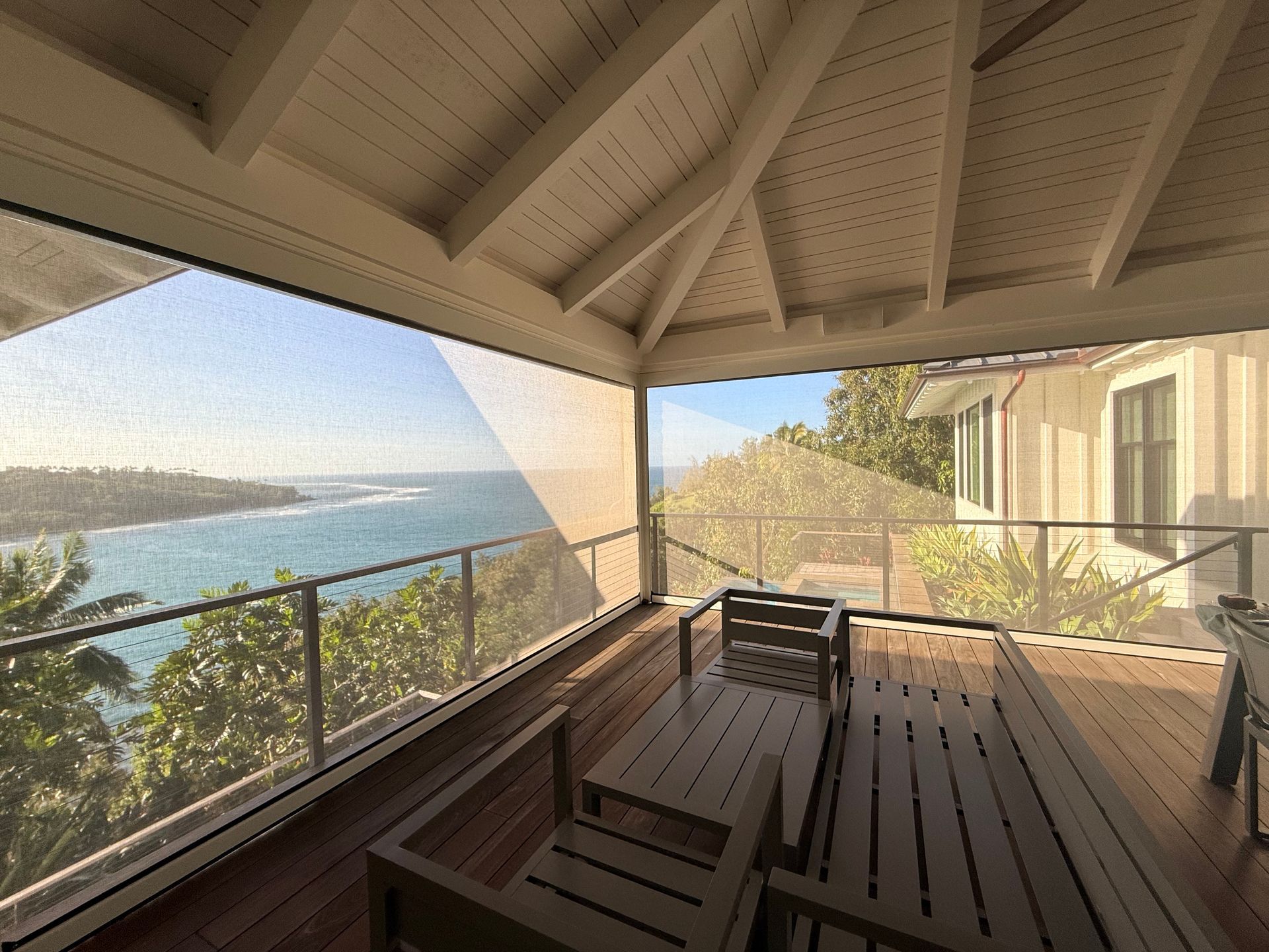 Covered wooden deck overlooking a blue ocean and coastline, with outdoor seating and a railing.