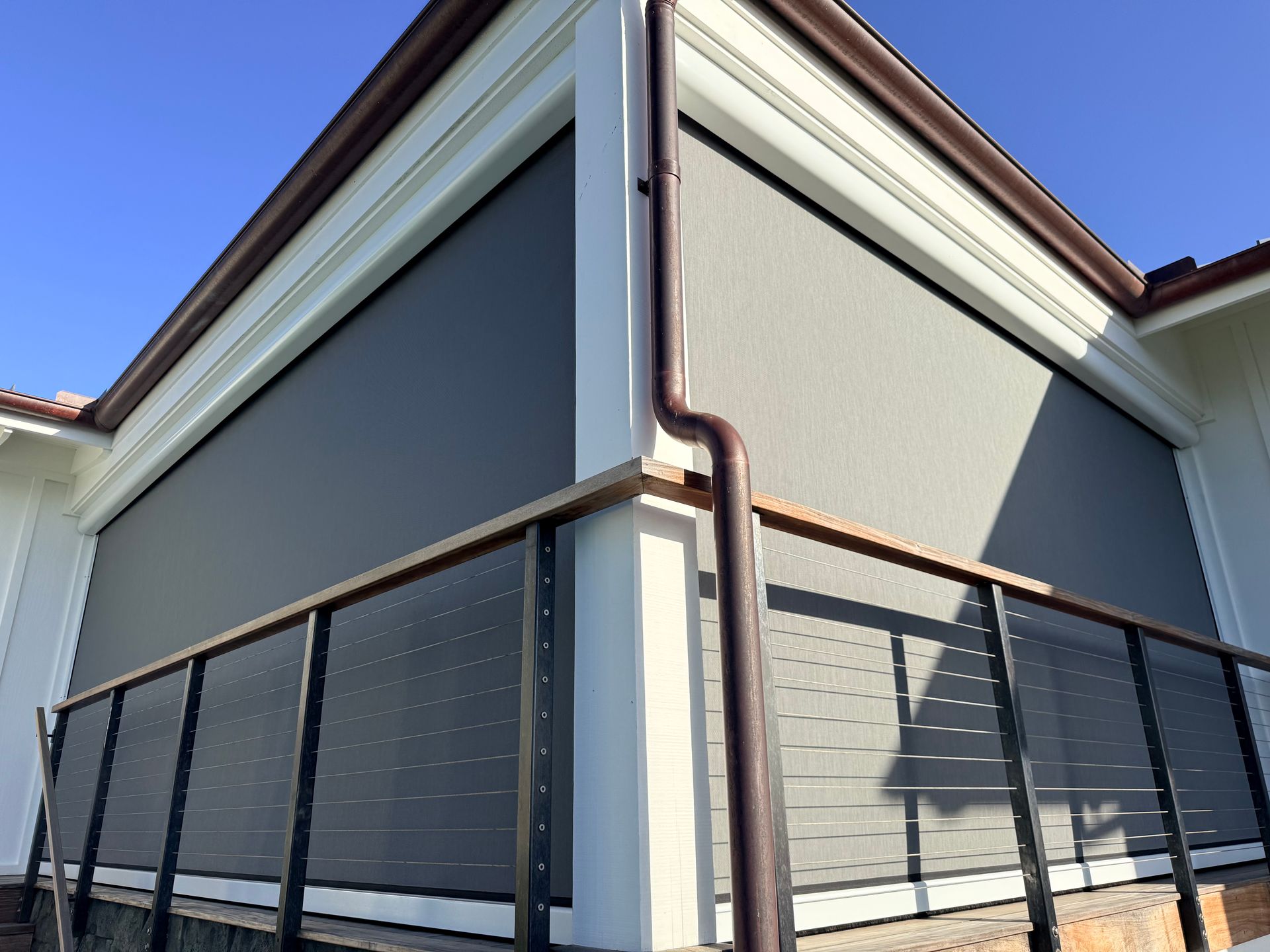 Corner of a modern building with gray roller shades and black balcony railing under a blue sky