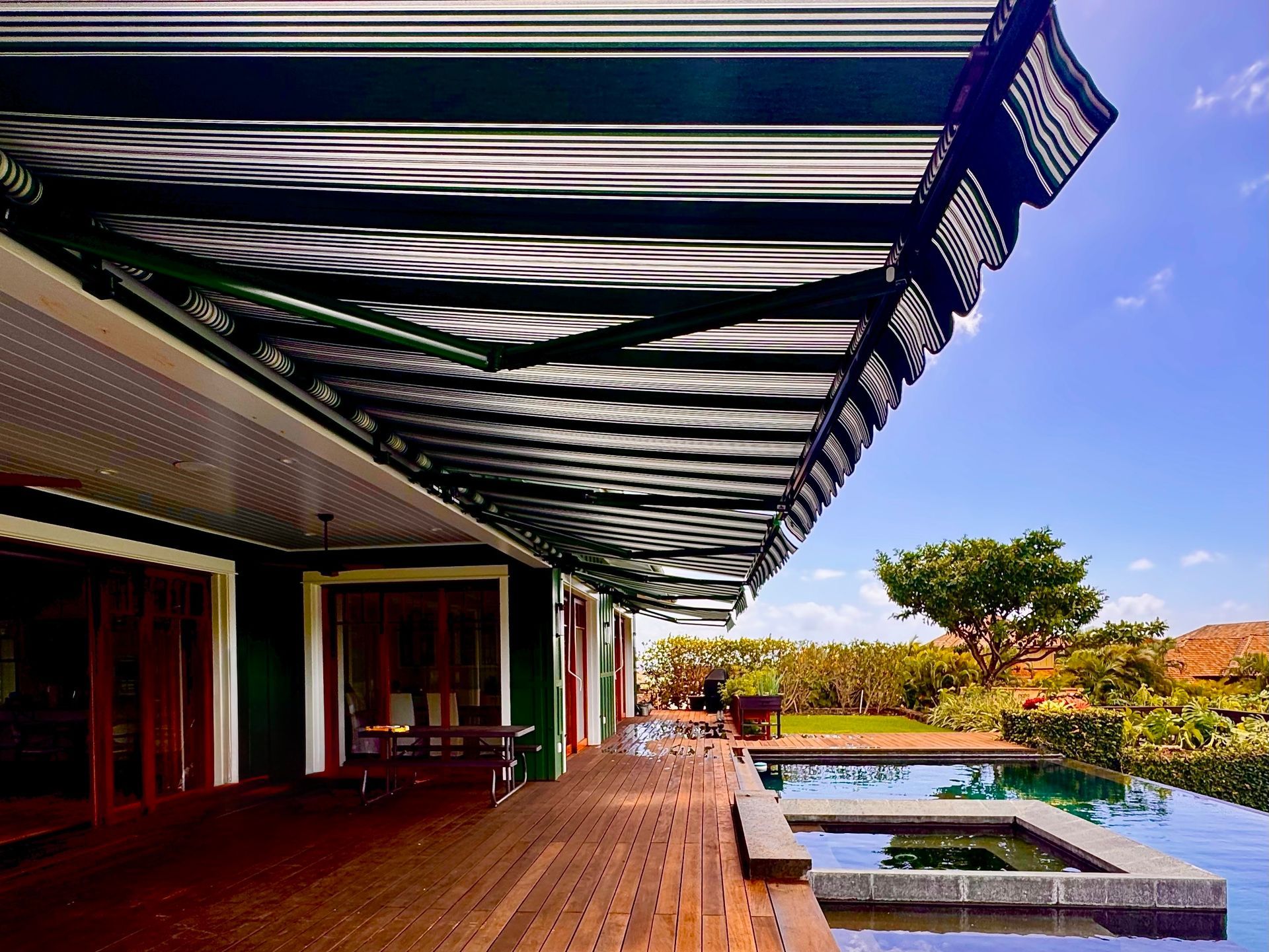 Modern patio with striped awning, pool, and garden under blue sky