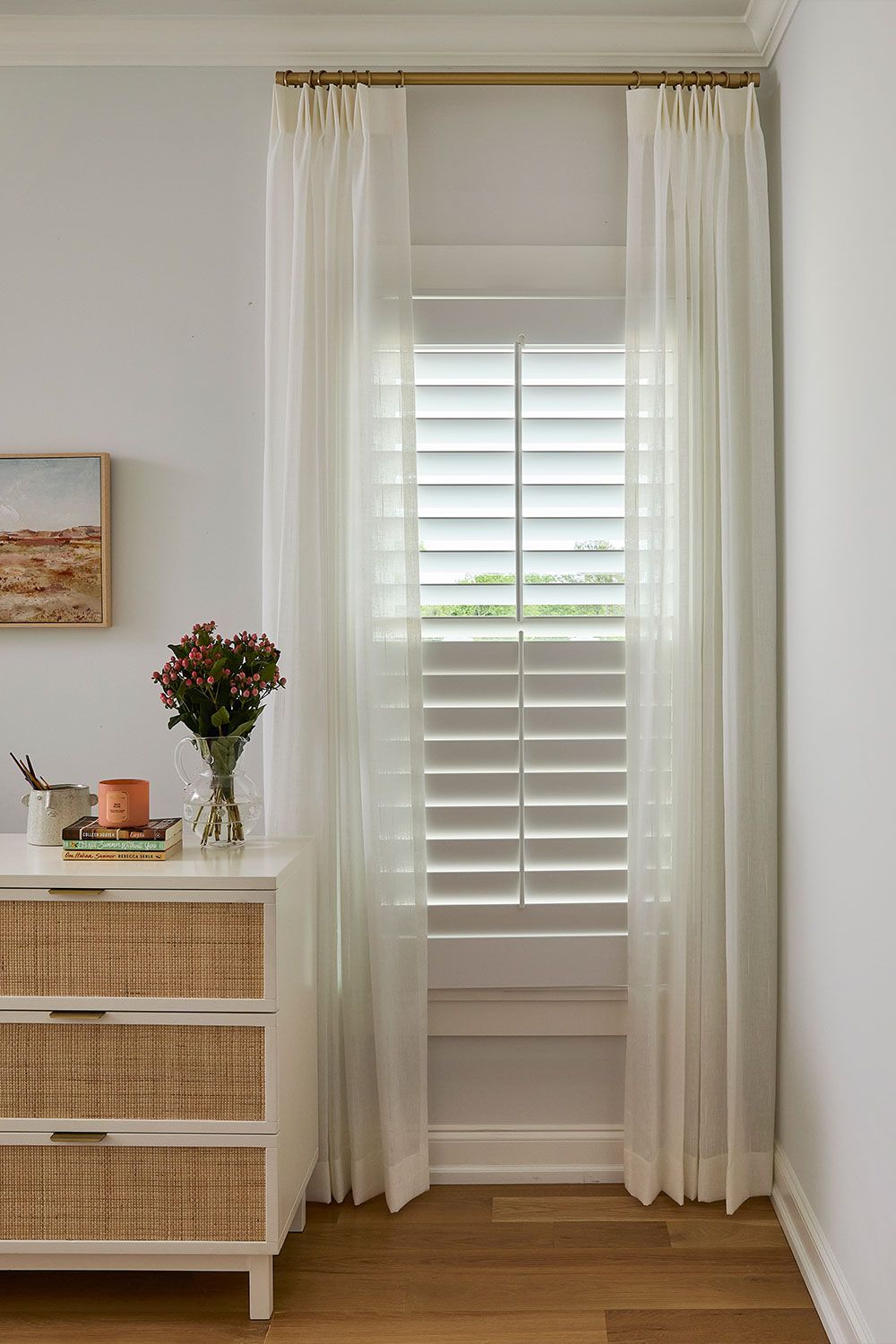 Bright bedroom corner with sheer curtains, white shutters, a woven dresser, and a vase of flowers