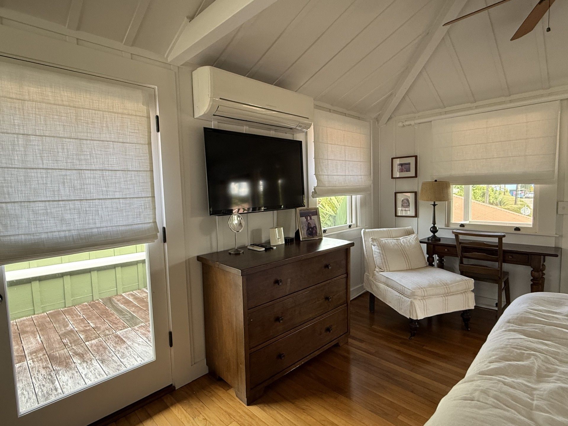 Bright bedroom with vaulted ceiling, TV over dresser, white chairs, hardwood floor, and windows with blinds.