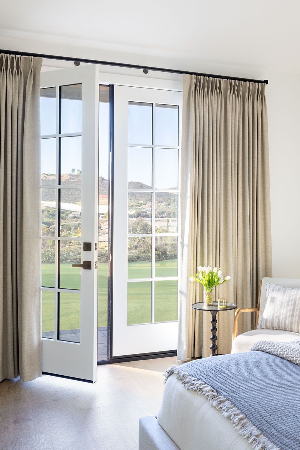 Bright bedroom with open French doors, beige curtains, and a bed by a sunny garden view