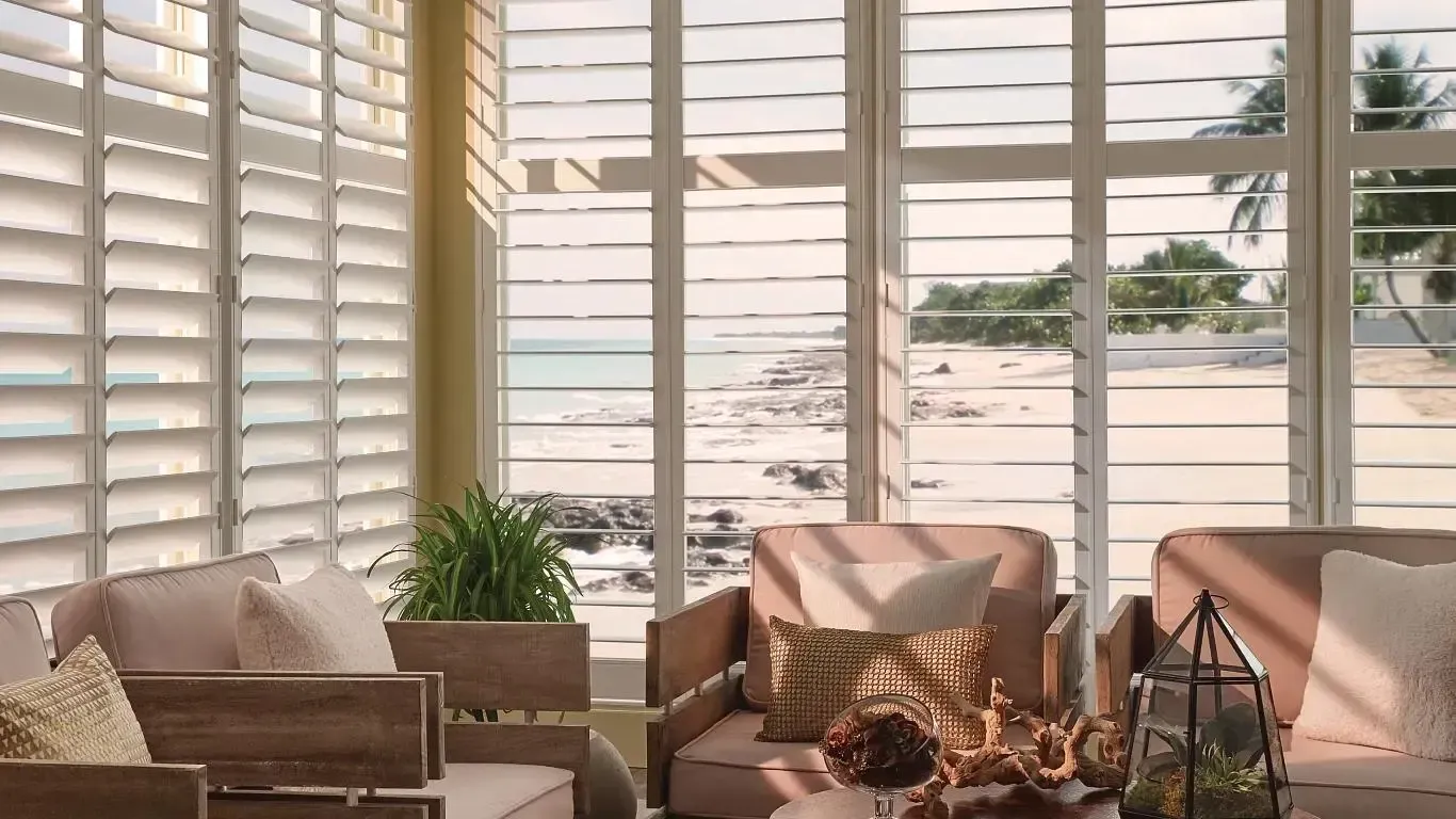 Sunlit living room with white shutters, beige sofas, and a potted plant by the window.