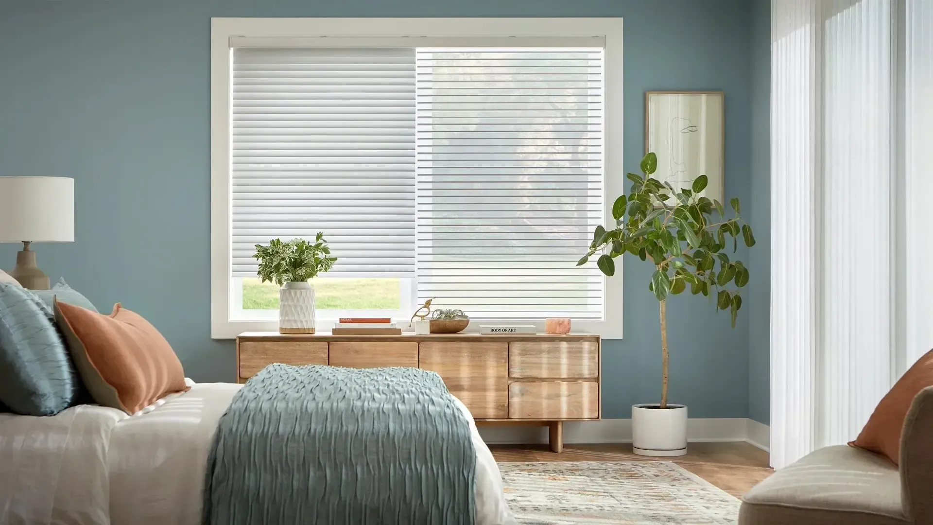 Bright bedroom with a bed, window blinds, dresser, lamp, and potted plants in soft neutral tones