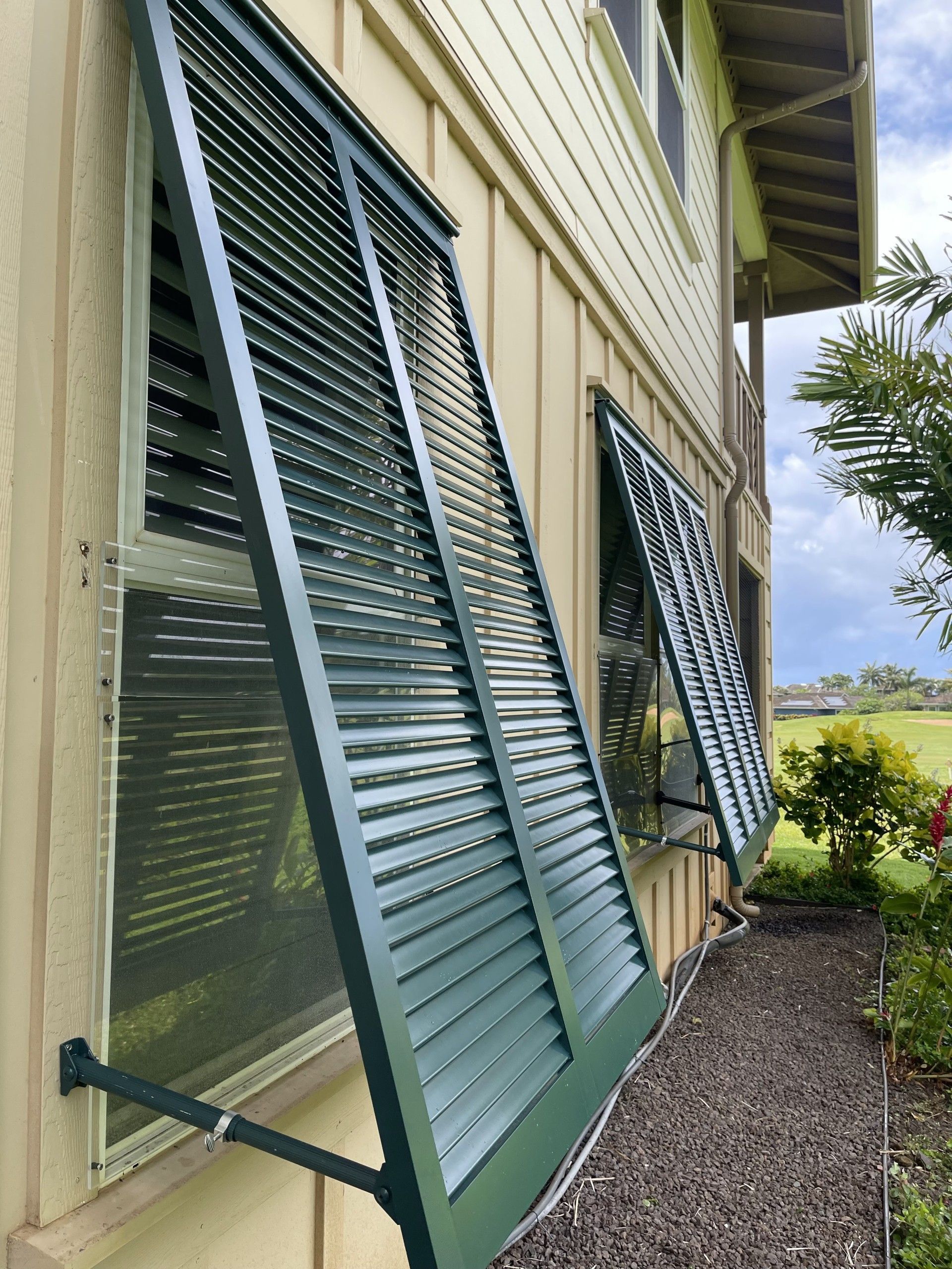 Green awning windows open on a cream house exterior above a gravel path.