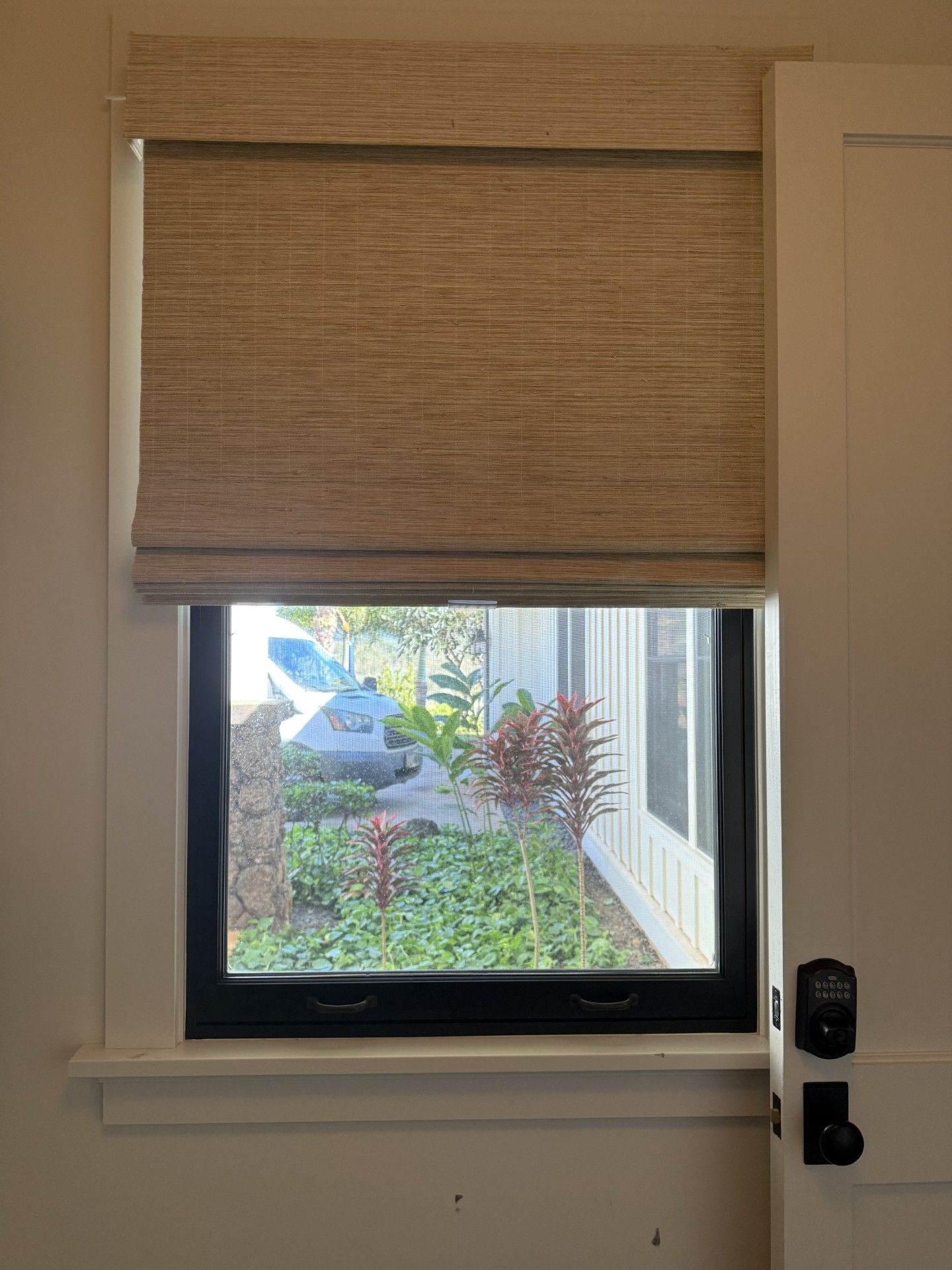 Small window with a beige roller shade partly down, showing a garden and house outside.