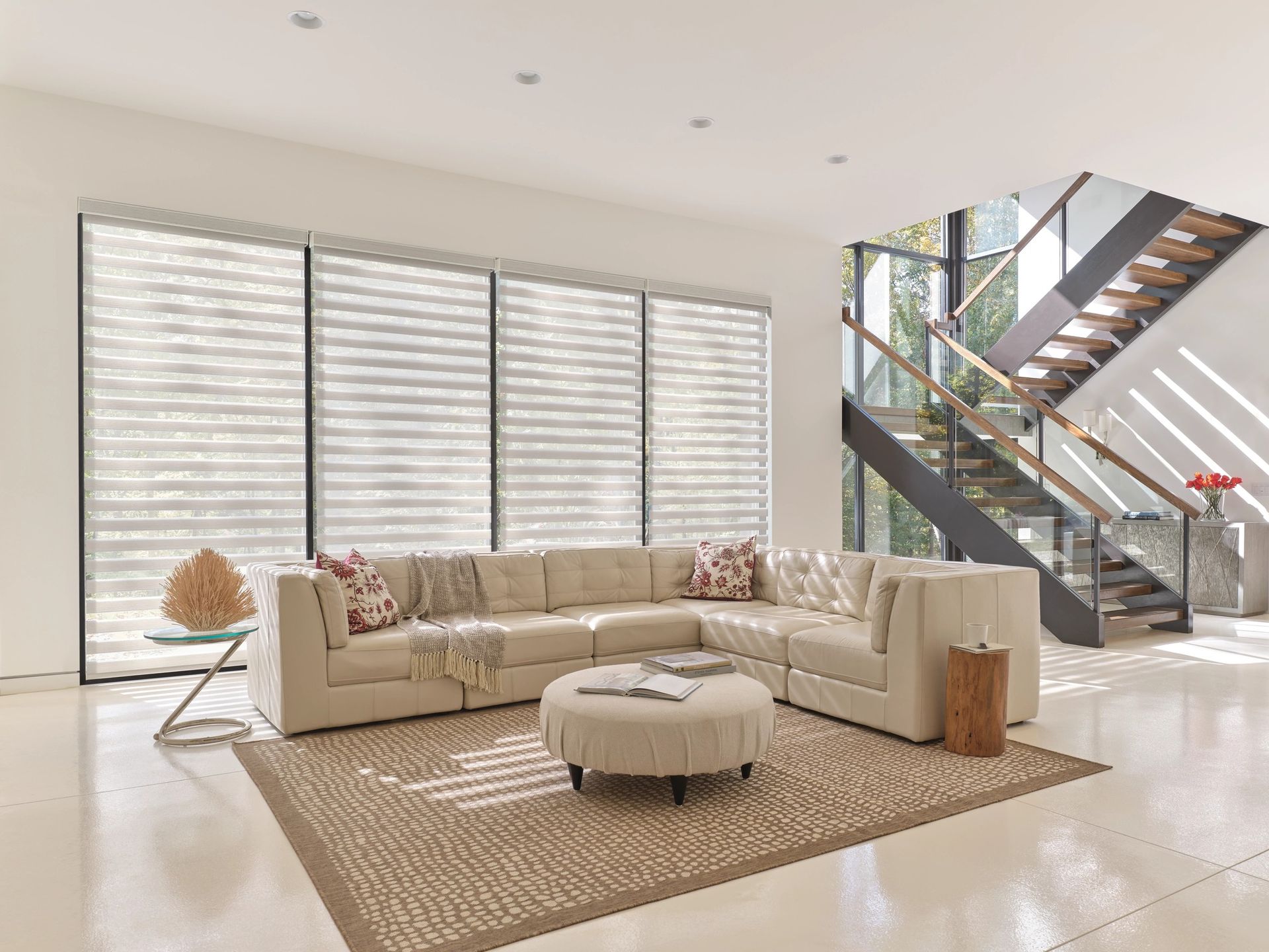 Bright modern living room with beige sectional, round ottoman, woven rug, and staircase by large windows
